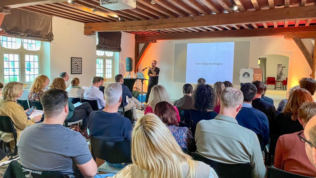 Een presentator staat op het podium voor een groep mensen in een bijeenkomst, met een slide die de tekst 'Eén miljard dingen' toont.